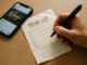 Hand holding a pen above a blank to-do list on a wooden desk with a phone displaying social media nearby.