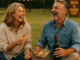 Middle-aged couple laughing together while enjoying tacos and wine at an outdoor picnic during golden hour. They sit barefoot on a striped blanket with string lights and a "Wine & Music Fest" sign in the background, creating a warm, joyful atmosphere.