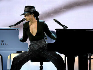 Alicia Keys performs on stage, seated between a black grand piano and a blue Yamaha piano, playing both at once. She wears a black hat, a plunging black top, and sequined pants.