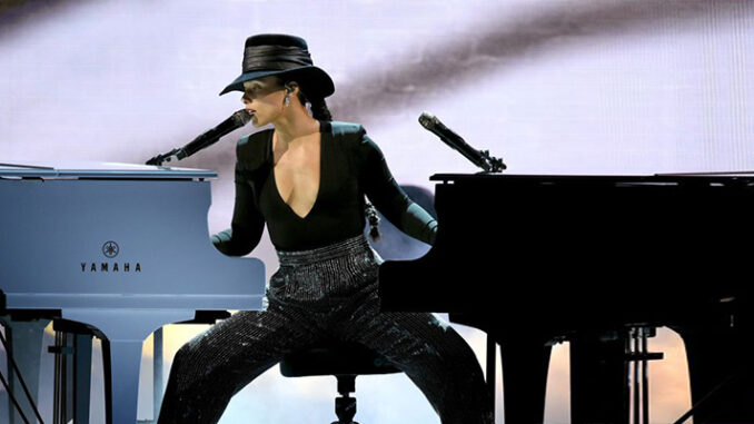 Alicia_Keys_Grammys_Living_Well_Magazine Alicia Keys performs on stage, seated between a black grand piano and a blue Yamaha piano, playing both at once. She wears a black hat, a plunging black top, and sequined pants.