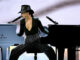 Alicia Keys performs on stage, seated between a black grand piano and a blue Yamaha piano, playing both at once. She wears a black hat, a plunging black top, and sequined pants.
