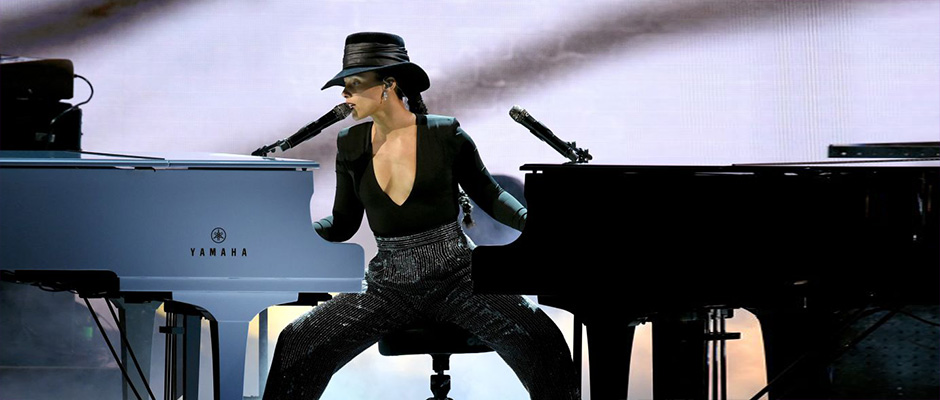 Alicia Keys performs on stage, seated between a black grand piano and a blue Yamaha piano, playing both at once. She wears a black hat, a plunging black top, and sequined pants.