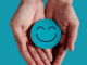 Two open hands gently hold a turquoise circle with a simple smiling face on it, symbolizing joy, calm, and positivity.