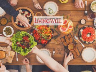 Overhead view of a vibrant shared meal with friends gathered around a wooden table. Colorful dishes include fresh salads, sliced tomatoes, olives, bread with dip, kabobs, and a pitcher of citrus water, creating a warm and inviting dining scene. The Living Well Magazine logo is placed at the center bottom.