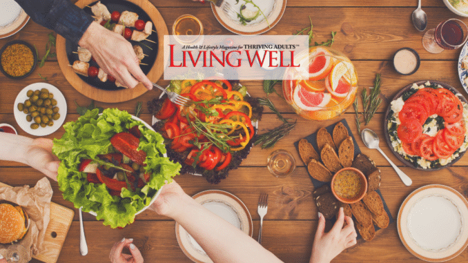 Gathering around the table Overhead view of a vibrant shared meal with friends gathered around a wooden table. Colorful dishes include fresh salads, sliced tomatoes, olives, bread with dip, kabobs, and a pitcher of citrus water, creating a warm and inviting dining scene. The Living Well Magazine logo is placed at the center bottom.