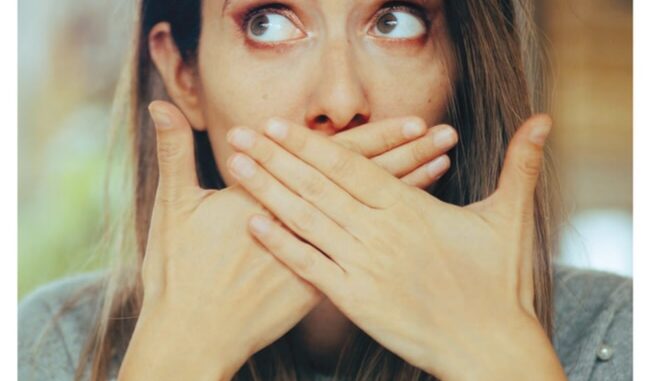 burping A surprised woman covers her mouth with both hands, looking upward with wide eyes, as if caught mid-burp.