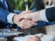 Two business professionals in suits shake hands across a polished desk, symbolizing trust and partnership during a legal consultation. A clipboard with papers and a pen rests in the foreground.