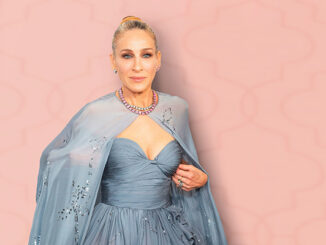 Sarah Jessica Parker poses elegantly in a strapless blue gown with a matching shawl and statement necklace against a soft pink patterned backdrop.