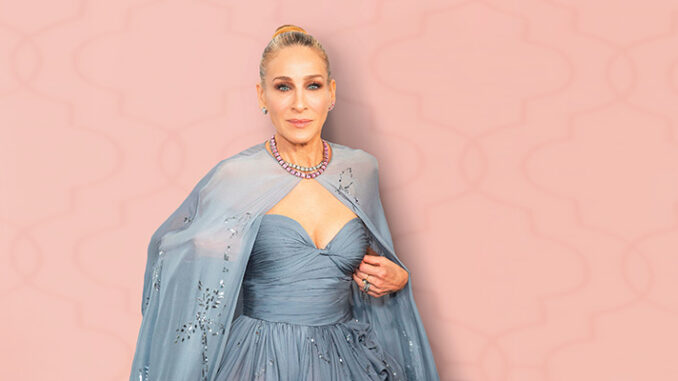 Sarah Jessica Parker poses elegantly in a strapless blue gown with a matching shawl and statement necklace against a soft pink patterned backdrop.