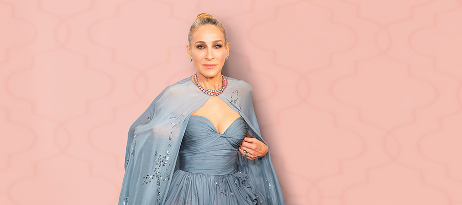 Sarah Jessica Parker poses elegantly in a strapless blue gown with a matching shawl and statement necklace against a soft pink patterned backdrop.