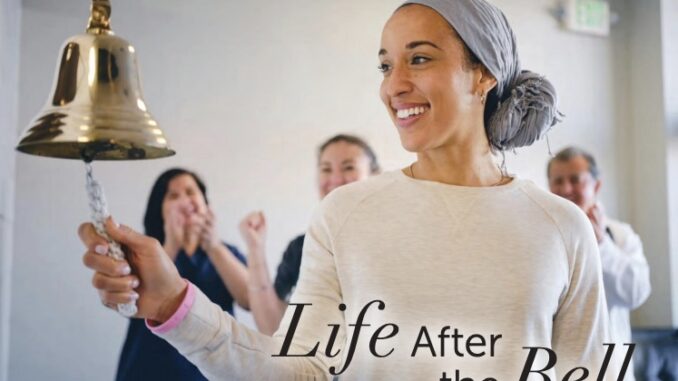 A smiling woman rings a large brass bell while loved ones cheer behind her, symbolizing the end of cancer treatment and the beginning of a hopeful new chapter in life.