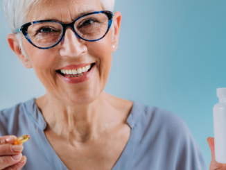 Smiling older woman with short white hair and glasses holding a fish oil capsule and a supplement bottle