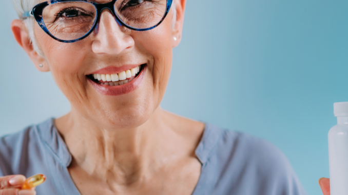 Smiling older woman with short white hair and glasses holding a fish oil capsule and a supplement bottle