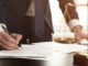 A lawyer in a suit signs a legal document at a desk beside the scales of justice